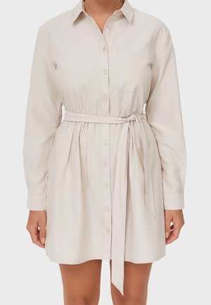 Kvinde iført en beige, langærmet skjortekjole med knapper, krave og bindebånd i taljen, stående mod en ensfarvet baggrund.