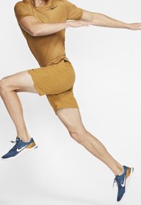 Nike Sportoutfit mit einem braunen Kurzarmshirt und passenden Shorts; marineblaue und orange Schuhe mit weißen Akzenten; Sprungpose.