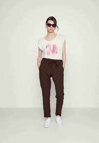 T-shirt branca com design abstrato rosa, calças castanhas ajustadas com cordão, e ténis brancos. Modelo usa óculos de sol pretos.