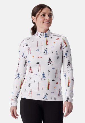 Femme portant un haut blanc zippé à manches longues avec des illustrations colorées sur le thème du ski, associé à un pantalon noir, souriante et regardant vers la droite.