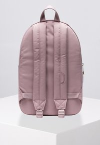 Mochila de tela rosa con correas acolchadas, textura suave y un asa en la parte superior. Cuenta con hebillas ajustables y un bolsillo lateral.