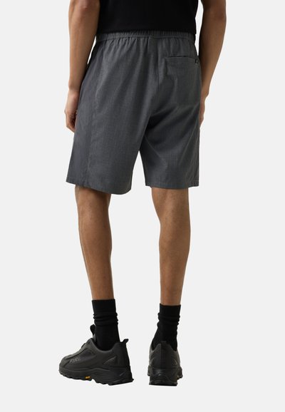 Mężczyzna ubrany w ciemnoszare krótkie spodenki do kolan, czarne skarpetki i czarne buty sportowe, pokazany od tyłu na jednolitym tle.