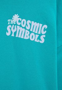 Tissu turquoise avec un texte rétro blanc disant « The Cosmic Symbols » et un petit dessin de fleur au-dessus du mot « The ».