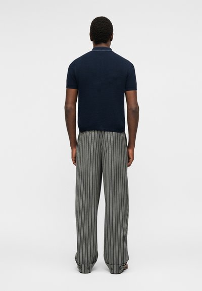 Homme de dos portant un polo en maille bleu marine à manches courtes et un pantalon gris à jambes larges avec des rayures verticales blanches.