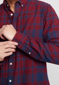 Rood en blauw geblokt flanel overhemd met een button-down kraag, witte plastic knopen en lange mouwen die bij de manchetten worden aangepast.
