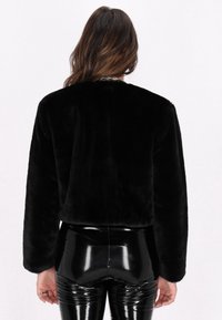 Schwarze Kunstfell-Shortjacke mit langen Ärmeln und glatter Textur. Kombiniert mit glänzenden, schwarzen, eng anliegenden Hosen.