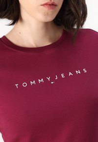 Burgundy puuvillane T-särk, millel on ümmargune kaelus ja lühikesed varrukad. Ülesehituse keskosas on valge "TOMMYJEANS" logo.