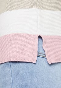 Maglione lavorato a maglia con strisce beige, bianche e rosa, caratterizzato da uno spacco sulla schiena, indossato sopra jeans di denim azzurro chiaro.