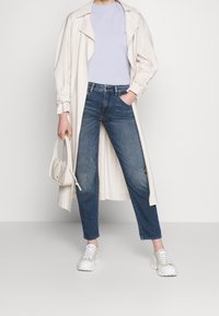 Abrigo trench beige claro sobre una camiseta de color púrpura claro, acompañado de jeans azules y zapatillas blancas. La bolsa tiene una superficie texturizada con un acabado suave.