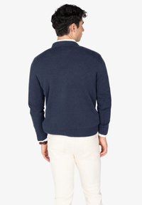 Suéter de punto azul marino con dobladillo y puños acanalados, con una textura sólida. Lleva puesto sobre una camisa blanca con cuello y pantalones de color crema.