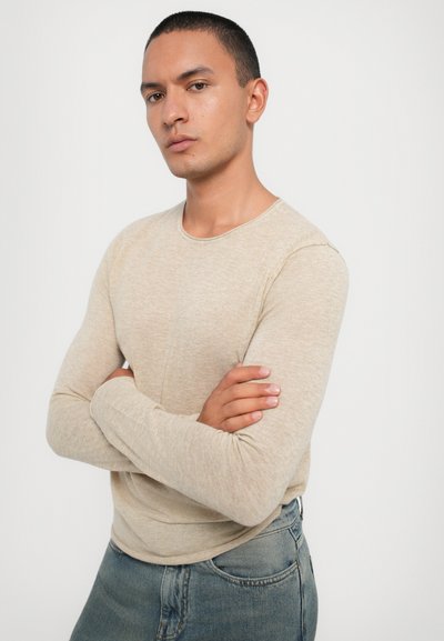 Beige pitkähihaine paita, joka on valmistettu kuvioidusta kankaasta ja jossa on pyöreä pääntie, yhdistettynä sinisiin farkkuihin. Malli seisoo kädet ristissä.