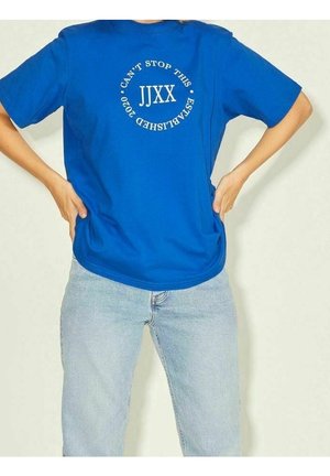 Person iført en blå T-shirt med hvid tekst "JJXX Can't Stop This Established 2020" og lyseblå jeans, hænderne hvilende på hofterne.