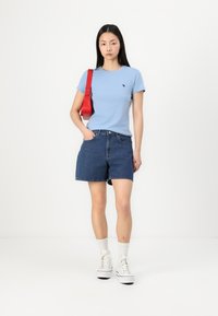 Light blue short-sleeve t-shirt, dark denim shorts, red shoulder bag, and white high-top sneakers with textured soles. Plain background.