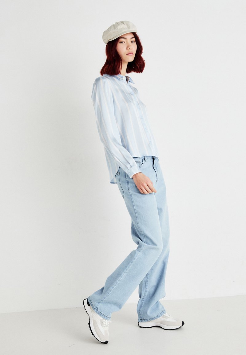 Camicia azzurra a righe, con bottoni, dalla vestibilità morbida, abbinata a jeans azzurri e sneakers bianche con suola nera; cappellino color crema.