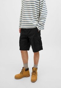 Czarne szorty cargo z wieloma kieszeniami, zestawione z brązowymi botkami do kostek oraz szarym, prążkowanym, długim rękawem, mającym luźny krój.