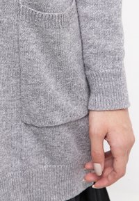 Suéter gris de punto con un bolsillo frontal, puños acanalados y una textura suave. La tela parece cálida y casual.