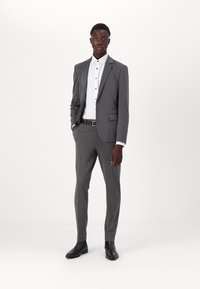 Costume gris ajusté avec un blazer à un bouton, un pantalon slim-fit et une chemise blanche à boutons noirs. Bottines en cuir noires.