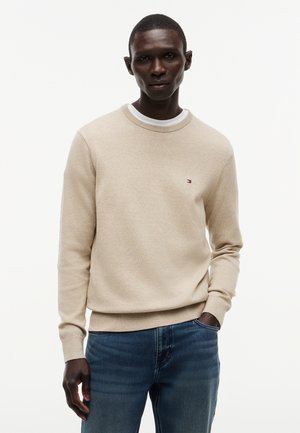 Mann trägt hellbeigen Pullover über weißem Hemd mit blauer Jeans, steht mit einer Hand in der Tasche vor einfachem Hintergrund.