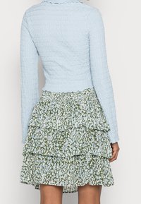 Femme portant un haut à manches longues texturé bleu clair et une jupe à motifs superposés verte et blanche, vue de dos.