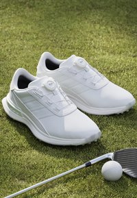 Bijele golf cipele imaju teksturirani gornji dio, BOA sustav vezivanja i tri bočne pruge. Postavljene na travu uz golf palicu i lopticu.