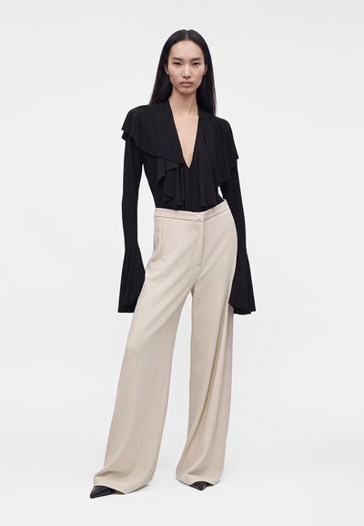 Femme portant un chemisier noir à manches longues avec volants et un col en V profond, et un pantalon large beige, debout sur un fond blanc.