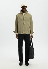 Homme portant une casquette beige, une veste vert olive, un pantalon noir et des chaussures noires, tenant un sac fourre-tout sombre sur un fond clair uni.