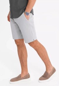 Shorts gris à la texture lisse, avec des poches latérales et de petites fentes au bas, associés à des chaussures brunes à enfiler et une chemise sombre.