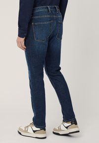 Jeans in denim slim-fit di colore blu scuro. Presentano cinque tasche, dettagli di cucitura e una vita di media altezza. Abbinati a sneakers beige e nere.