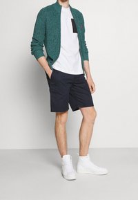 Teal stickad zip-up cardigan över en vit t-shirt med en svart ficka, kombinerat med marinblå shorts och vita high-top sneakers.