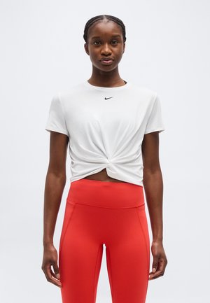 Modèle féminin debout, portant un haut blanc à manches courtes noué avec un petit logo Nike noir et des leggings rouges taille haute.
