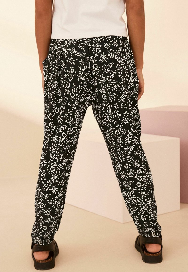 Personne portant un pantalon large à motif floral noir et blanc et des sandales noires, debout les mains dans les poches contre un fond neutre.