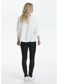 Chemisier blanc à manches bouffantes et poignets texturés, assorti à un pantalon noir ajusté et des baskets blanches. Design épuré, détails minimaux.