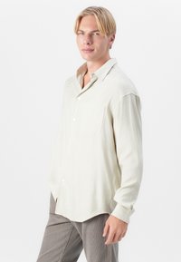 Lys beige langermet skjorte laget av teksturert stoff, med knapping foran og en enkelt brystlomme. Kombinert med lysegrå bukser.
