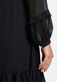 Schwarzes Kleid mit transparenten, plissierten langen Ärmeln und einem gerüschten Saum. Der Stoff ist leicht und strukturiert. Knopfdetails an den Manschetten.