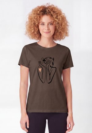 Jonge vrouw met krullend haar die een bruin t-shirt draagt met een zwart lijntekening van een persoon die een bloem vasthoudt en een klein hartje op de arm.