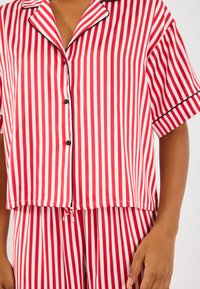 Red and white striped pajama set featuring short sleeves, a button placket, and drawstring waist. Smooth fabric with contrasting black accents.