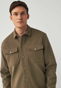 Camisa marrón de botones con una tela texturizada, que presenta un cuello con solapa, dos bolsillos en el pecho y botones de presión metálicos, exhibida sobre un fondo neutro.