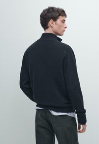 Maglione lavorato a maglia grigio scuro con colletto alto, polsini e orlo a costine, caratterizzato da una vestibilità rilassata e un tessuto testurizzato. Indossato su una camicia bianca.