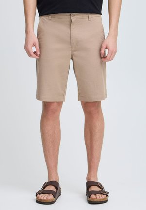Beige shorts in a straight-cut design, made of lightweight material; features front pockets and a button closure. Worn with brown sandals.