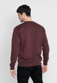 Mand med kort mørkt hår iført en vinrød langærmet sweatshirt og sorte bukser, vendt væk mod en ensfarvet lys baggrund.