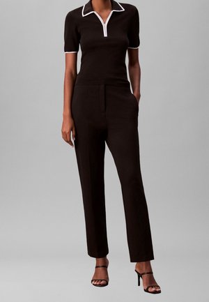 Femme portant un haut noir à manches courtes avec col et bordures blanches, un pantalon noir taille haute, et des sandales à talons hauts noires à lanières, debout.