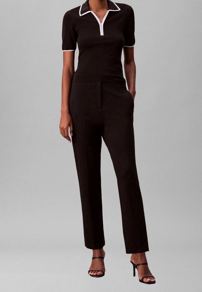 Femme portant un haut noir à manches courtes avec col et bordures blanches, un pantalon noir taille haute, et des sandales à talons hauts noires à lanières, debout.
