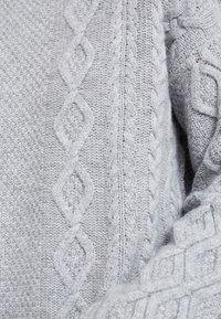 Světle šedý pletený svetr s texturovanými vzory, diagonálními kabely a diamantovými tvary, vyrobený z měkkého materiálu připomínajícího vlnu.