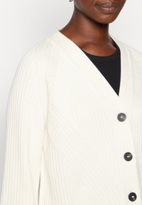 Krämfärgad ribbad cardigan med v-ringning, prydd med fem svarta knappar och ett texturerat mönster, tillverkad av mjukt material för komfort.