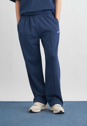 Osoba v tmavomodrých voľných teplákoch s rukami v vreckách a béžovými teniskami stojaca na modrom koberci pred sivým pozadím.