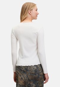 Langärmlige, weiße Bluse mit einem geschwungenen Ausschnitt und Saum. Der Stoff ist glatt, figurnah und wird mit einem gemusterten Rock kombiniert.