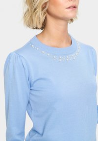 Maglione azzurro chiaro in maglia a coste, con maniche a sbuffo e perline decorative attorno al collo, che offrono un dettaglio testurizzato.