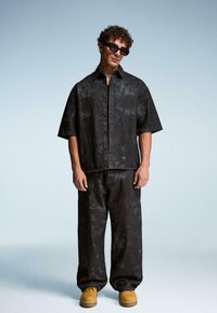 Uomo che indossa una camicia a maniche corte larga con un motivo scuro e pantaloni abbinati, con stivali beige e occhiali da sole neri rettangolari, in piedi contro uno sfondo semplice.