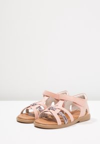 Różowe sandałki dla małych dzieci z błyszczącymi paskami, regulowanymi zapięciami na rzep i miękką brązową podeszwą na białej powierzchni.