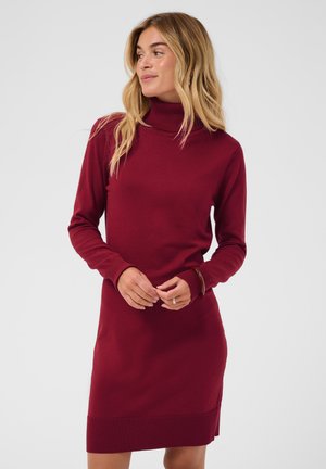 Femme aux longs cheveux blonds portant une robe-pull à col roulé rouge, debout devant un fond clair uni.